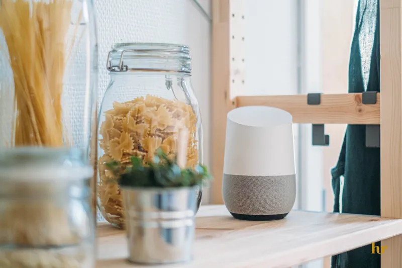 Voice assistant smart speaker on a wooden kitchen shelf next to a glass jar of bow-tie pasta