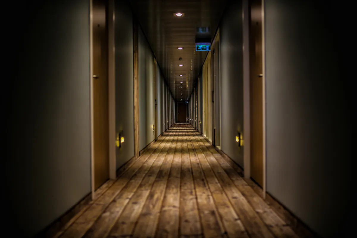 Dimly lit hotel corridor with a long wooden floor, rows of guest room doors and a glowing exit sign