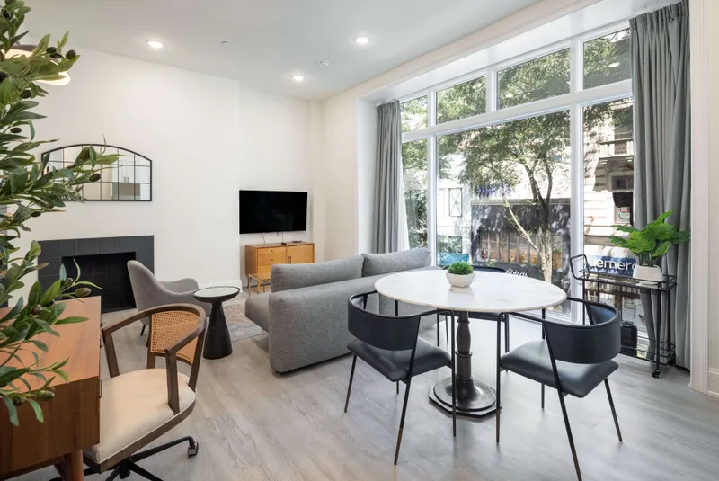 Modern apartment living room with a gray sofa, round marble dining table and a tall window onto street trees