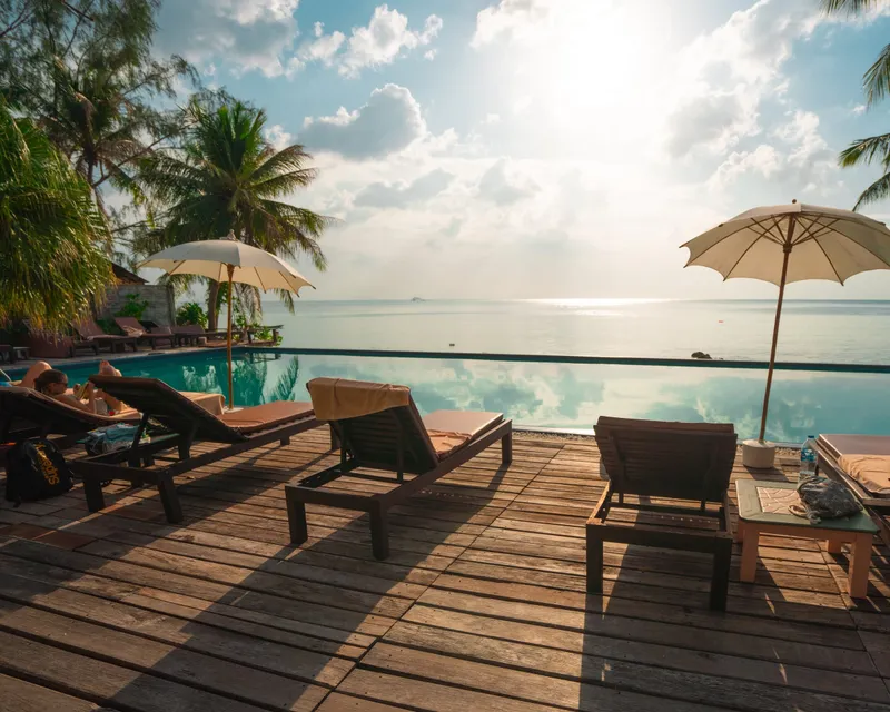 Wooden pool deck with lounge chairs and cream umbrellas beside an infinity pool facing the sea at sunset