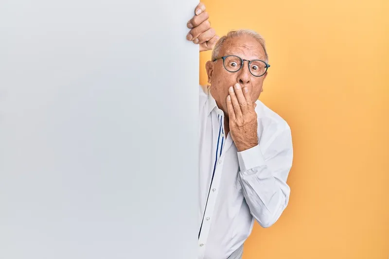 Surprised older man in glasses peeking from behind a blank white panel with a hand over his mouth, yellow background