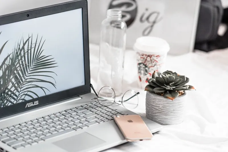 Laptop, glasses, phone, water bottle, coffee cup and a small succulent arranged on a white bedsheet