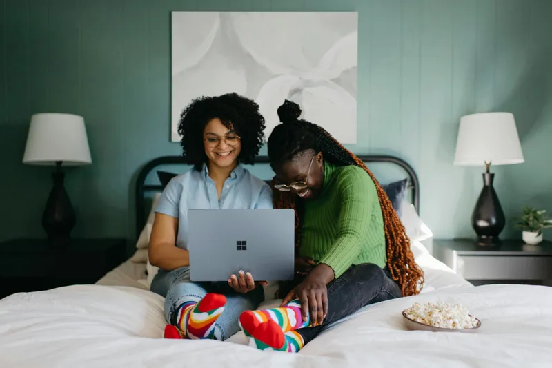 Two women sitting on a bed in a teal guest room, laughing at a laptop with a bowl of popcorn beside them